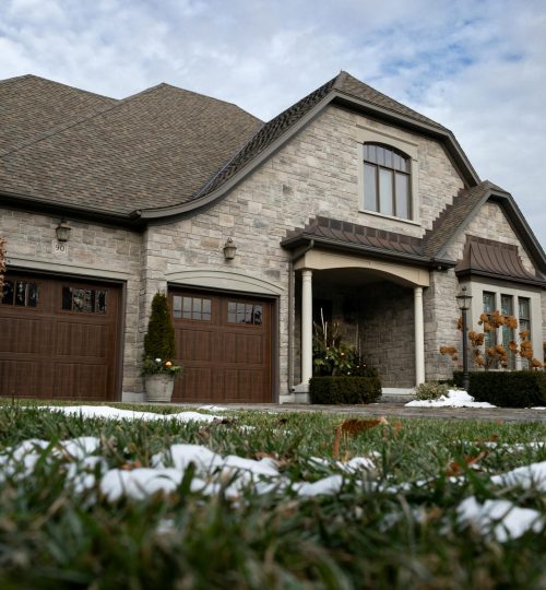 Projects Charming stone house with wooden garage doors and a snow-dusted lawn under a cloudy winter sky.