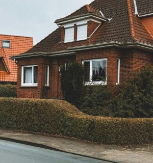 ASIGURARI LOCUINTA A cozy red brick house with a sloped roof and trimmed hedges in a suburban neighborhood.