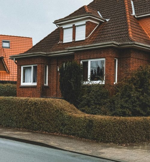 Projects A cozy red brick house with a sloped roof and trimmed hedges in a suburban neighborhood.